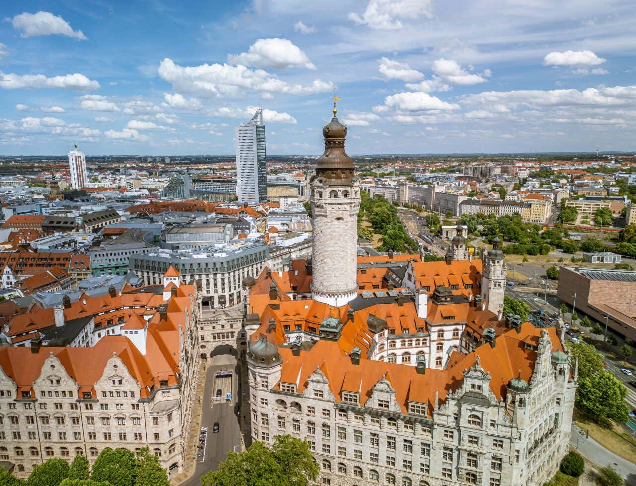 An aerial perspective captures the sprawling, red-roofed New Town Hall of Leipzig with its massive central stone tower standing prominently against the city's modern urban skyline.