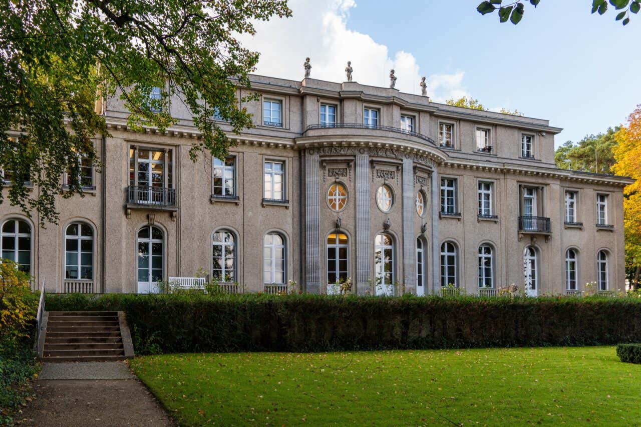 The grand, multi-story facade of the House of the Wannsee Conference stands behind a manicured green lawn and a dark hedge under a bright, partly cloudy sky.