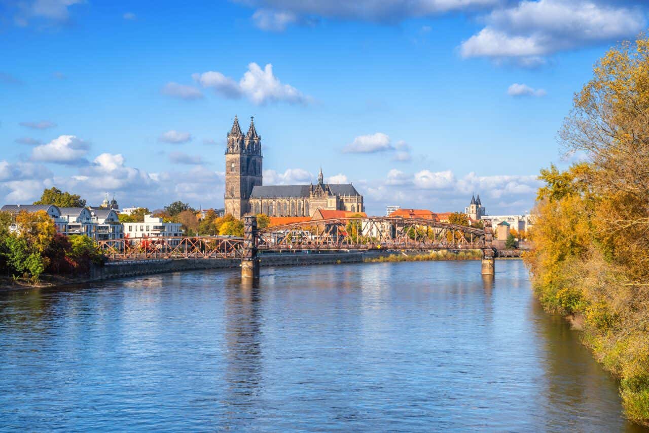 The towering gothic Magdeburg Cathedral and a historic iron bridge are reflected in the calm waters of the Elbe River, framed by golden autumn trees on the riverbank.