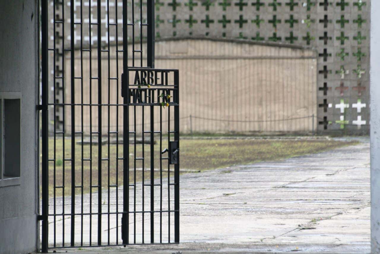 The iron gate at the entrance of the Sachsenhausen concentration camp memorial, featuring the infamous inscription "Arbeit Macht Frei" set against a backdrop of a gravel courtyard and a concrete wall with cross-shaped cutouts.