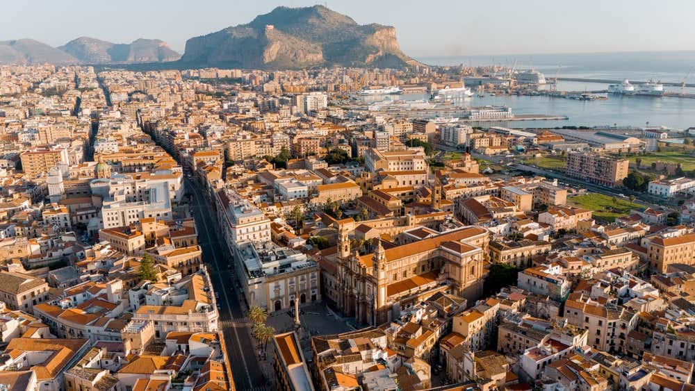Vista aerea di Palermo con il centro storico in primo piano, con edifici storici dai tetti arancioni. Sullo sfondo si erge il Monte Pellegrino, la montagna che domina la città, mentre sulla destra si intravede il porto e il Mar Tirreno. L'immagine cattura la densità urbana del capoluogo siciliano e il suo rapporto tra mare e montagna