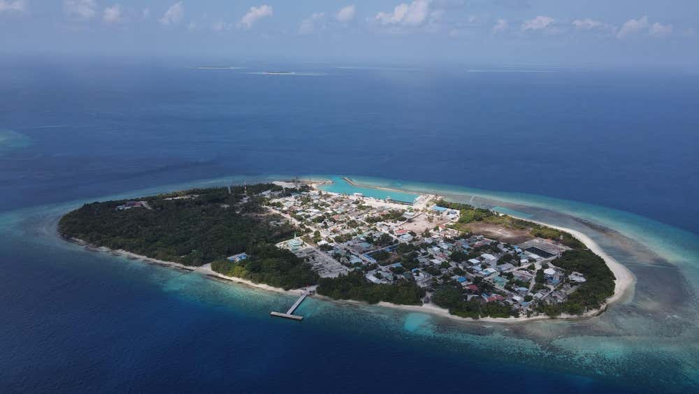 Vista aérea panorámica del Atolón de Raa en Maldivas, con una exuberante isla tropical cubierta de vegetación y núcleos habitados, rodeada de arrecife de coral y aguas turquesas del océano Índico