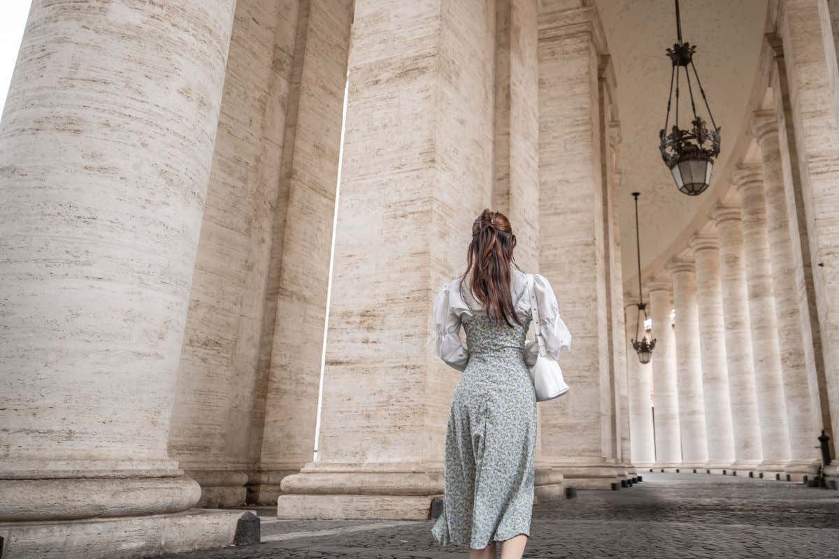 Una mujer de espaldas a la cámara caminando entre un pasadizo de columnatas monumentales pertenecientes a la plaza de San Pedro en el Vaticano