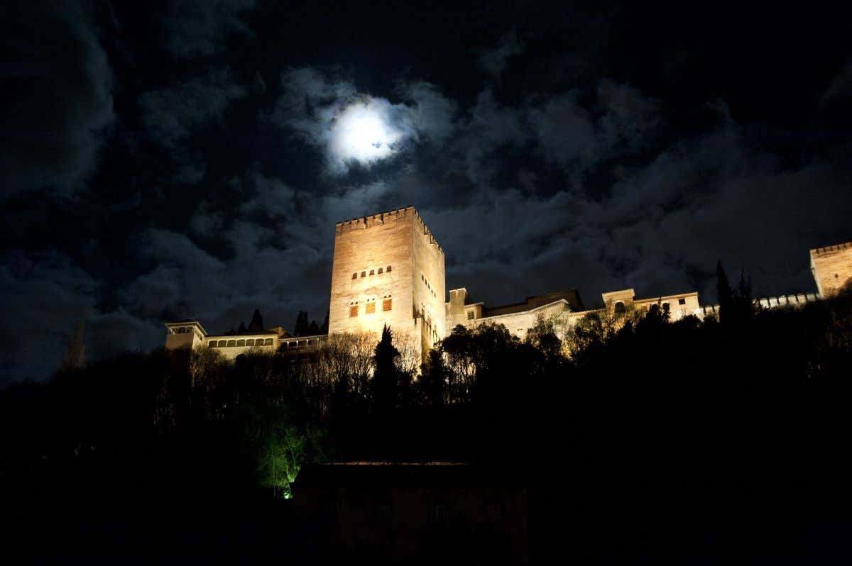 Alhambra de Granada iluminada al caer la noche con la luna de fondo cubierta por las nubes