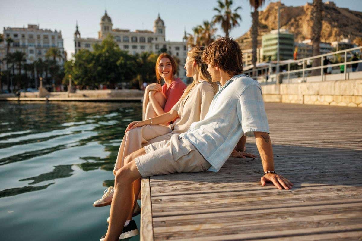 Tres jóvenes en verano sentados en la orilla del puerto de Alicante con la ciudad al fondo
