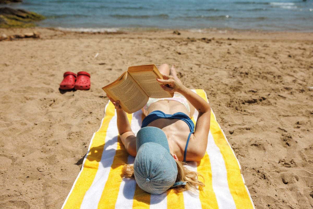 Una mujer tumbada en la playa de la Comunitat Valenciana en Semana Santa en una toalla de rayas amarillas y blancas leyendo un libro