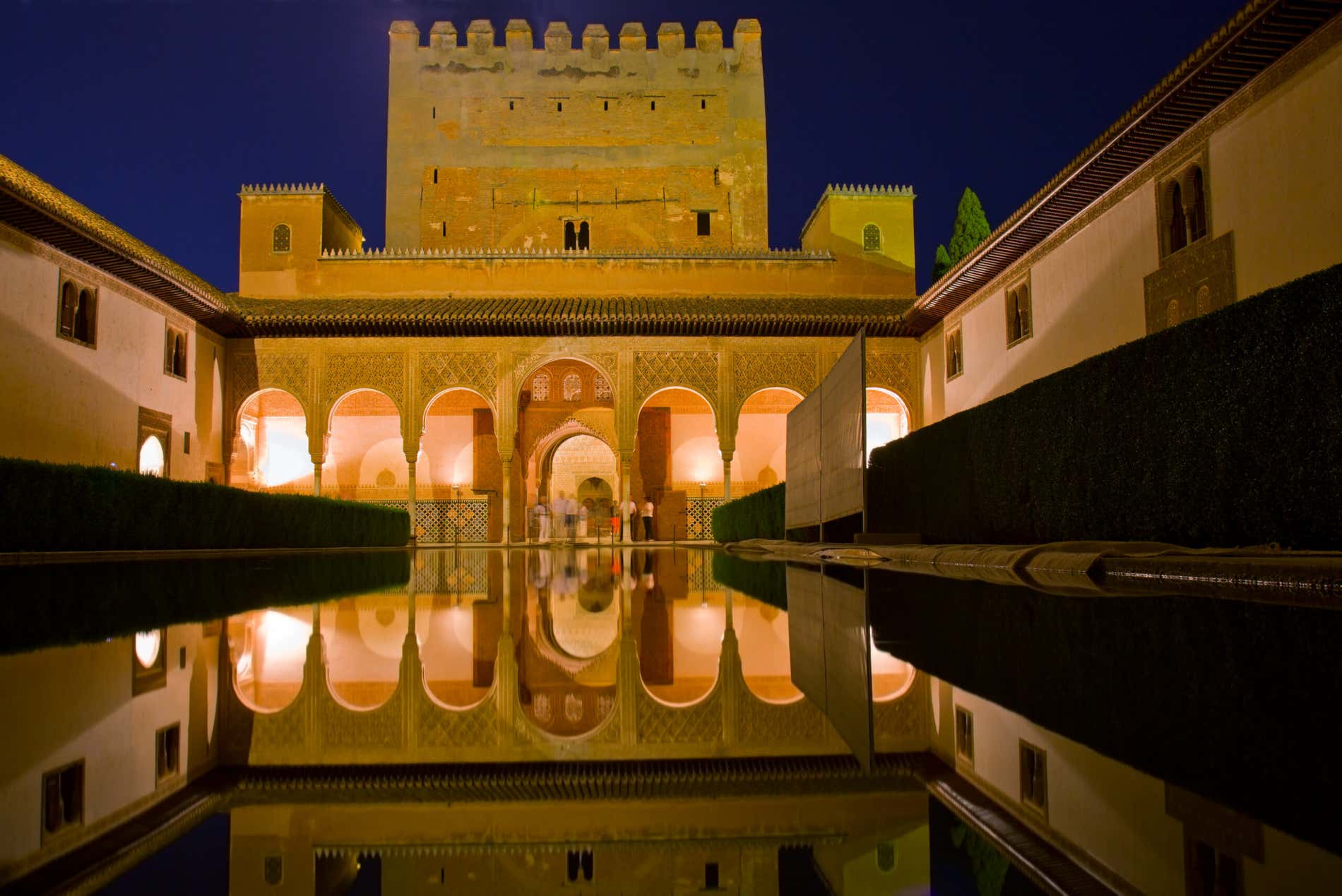 Cortile dell'Alhambra di Granada di notte, con una parte del Palazzo illuminata che si specchia nell'acqua e alcune persone sullo sfondo