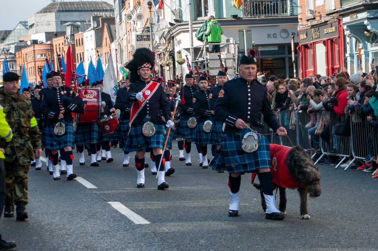 Men wearing kilts holding instruments and a dog parade down a street in Dublin at Easter as people look on.