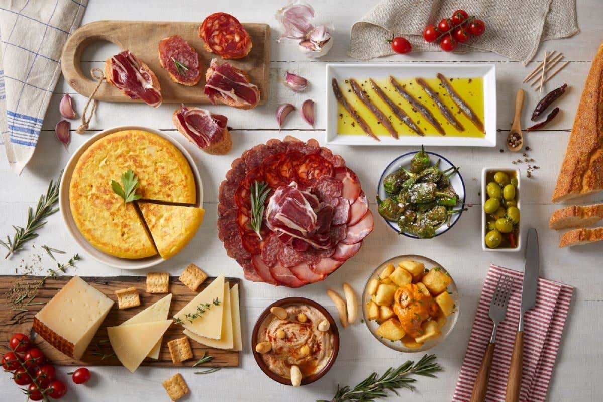 Plates of tortilla, cured meats, padrón peppers, olives, cheese boards, patatas bravas, and hummus.
