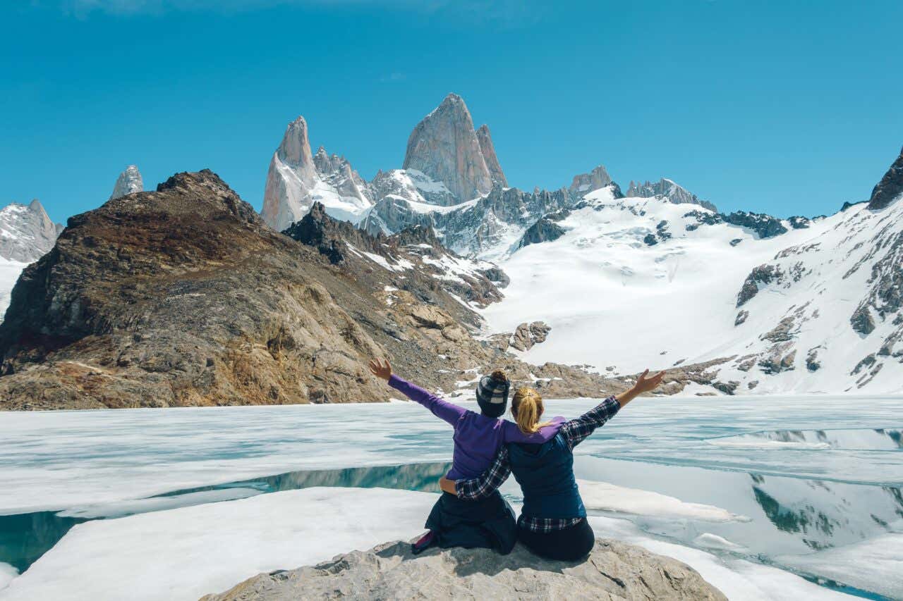 Due donne abbracciate di spalle e con un braccio in aria osservano montagne rocciose innevate
