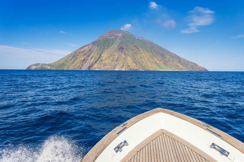 La prua di una barca mentre naviga verso l'isola vulcano di Stromboli in un giorno di sole, mentre dalla cima dell'isola escono degli sbuffi bianchi di fumo