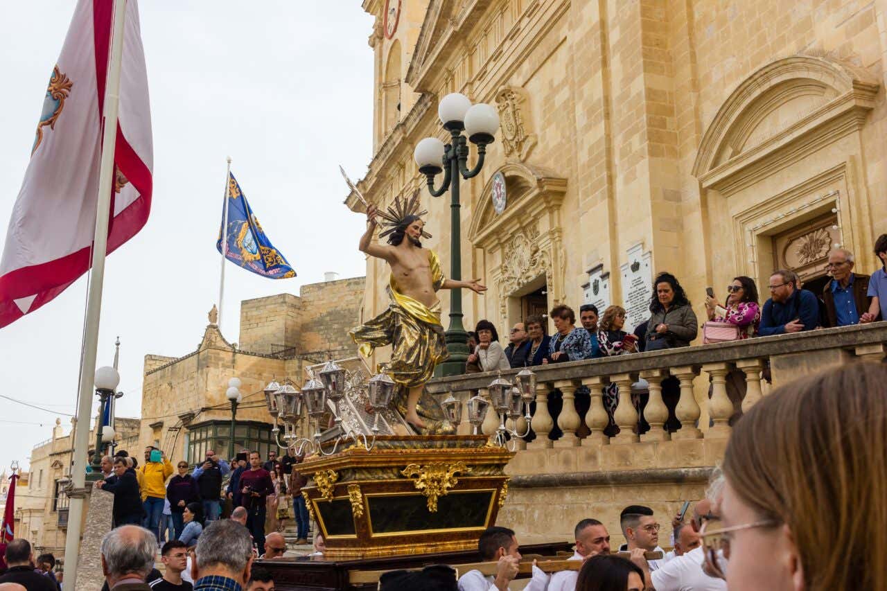 Easter procession with a Jesus statue and people looking on in front of a large monumental building in Malta.