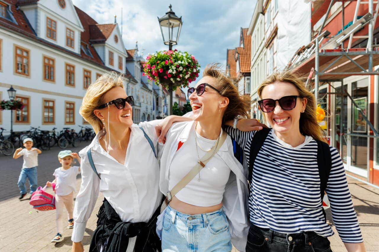 Three women hug and smile as they walk down a European pedestrian street.