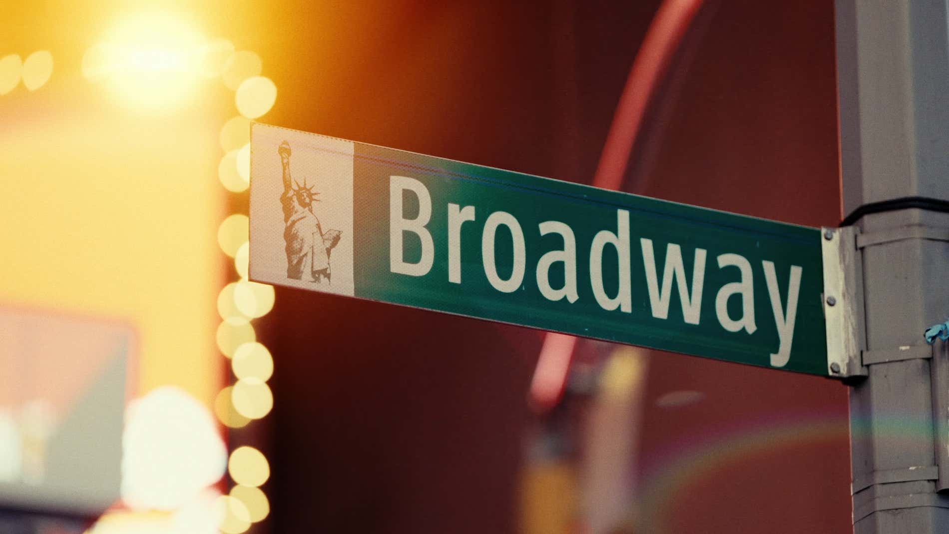 Close-up da placa da rua Broadway com a Estátua da Liberdade ao fundo, localizada em Manhattan, Nova York