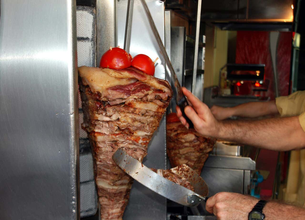 A Gyros meat spit spinning in a kitchen with a chef holding knives.