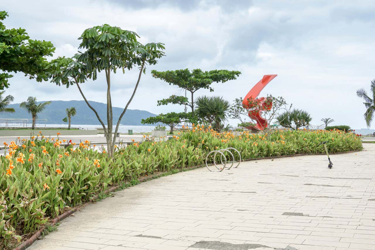 Um calçadão com jardins floridos e vista para a icônica escultura vermelha de Tomie Ohtake