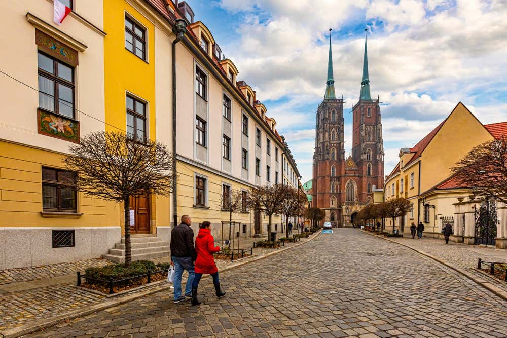 Una strada in ciottoli a Breslavia (Wrocław) affiancata da edifici storici colorati in toni di giallo e crema, con una coppia che cammina in primo piano e le due alte torri della Cattedrale di San Giovanni Battista che svettano sullo sfondo