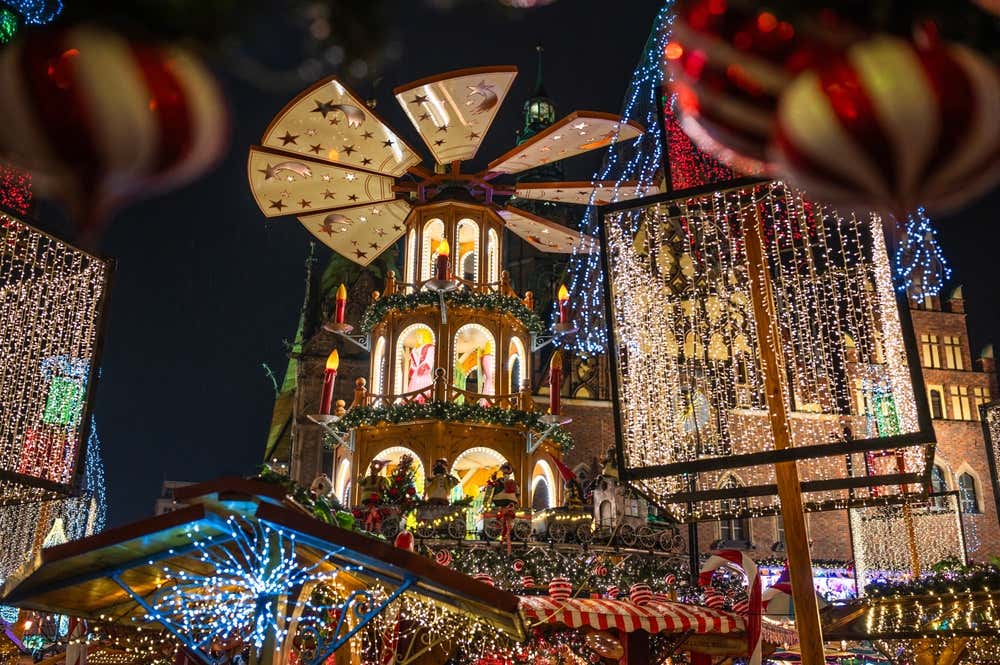 Una grande piramide natalizia in legno illuminata al centro di un mercatino di Natale notturno, circondata da luminarie brillanti, decorazioni sospese e bancarelle addobbate