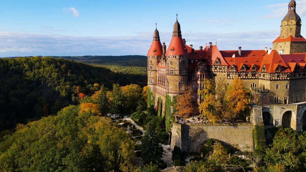 Vista panoramica dall'alto del Castello di Książ in Polonia, un imponente palazzo fiabesco con tetti rossi e torri appuntite, circondato da rigogliose foreste con colori autunnali e giardini terrazzati