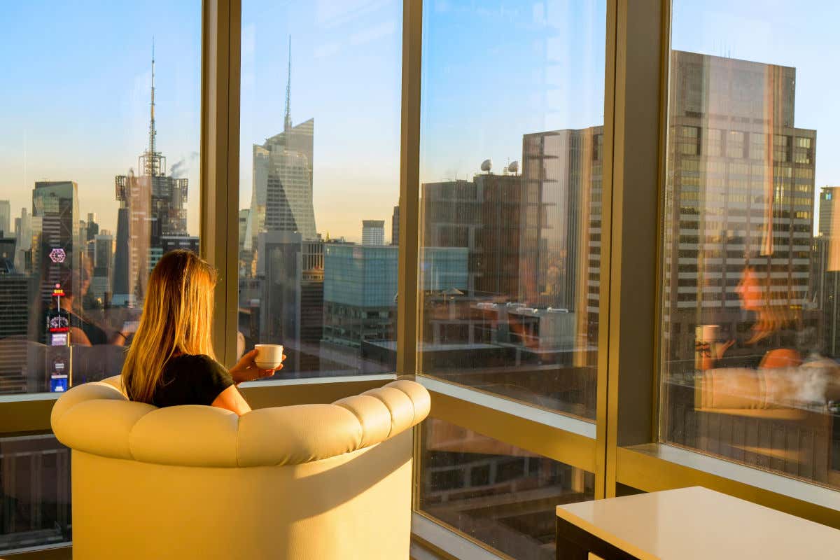 Una turista de espaldas a la cámara sentada y tomando una taza de café junto a las ventanas de un rascacielos con vistas a otros edificios de Nueva York