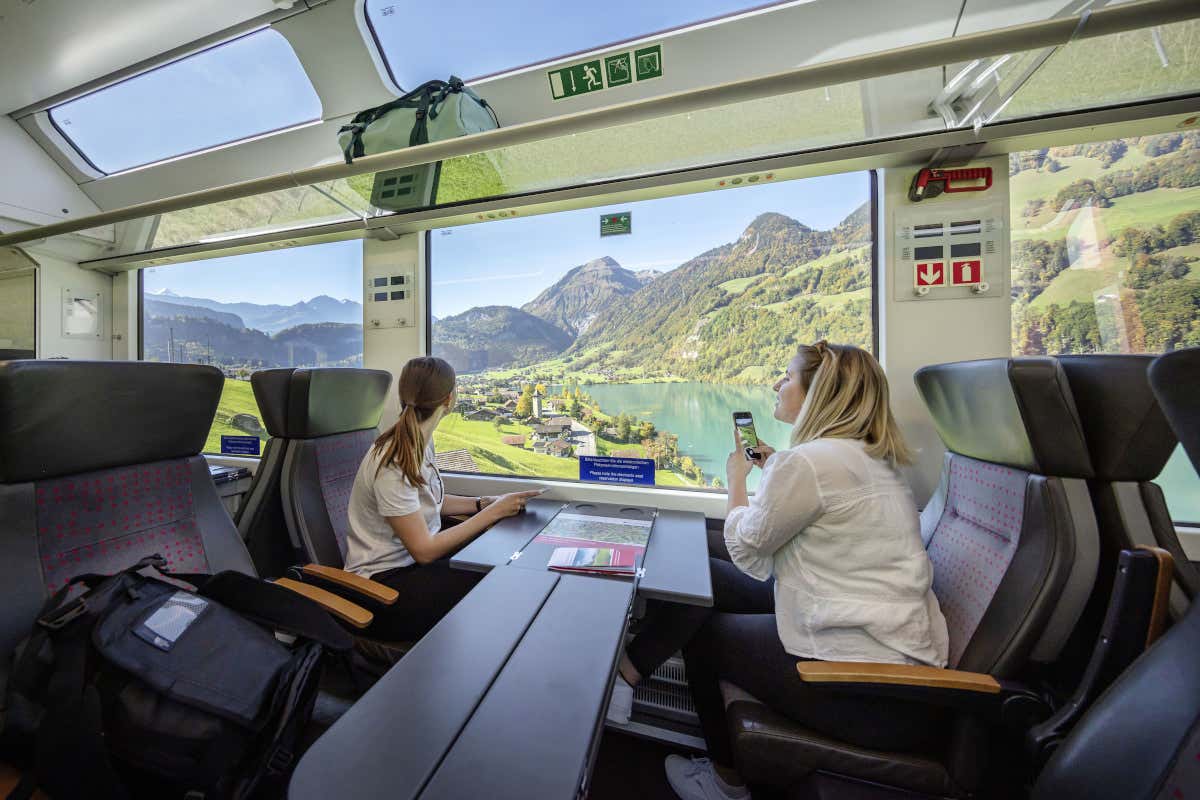 Dos mujeres hacen fotografías panorámicas de un verde paisaje de lagos y montañas desde el interior de un tren