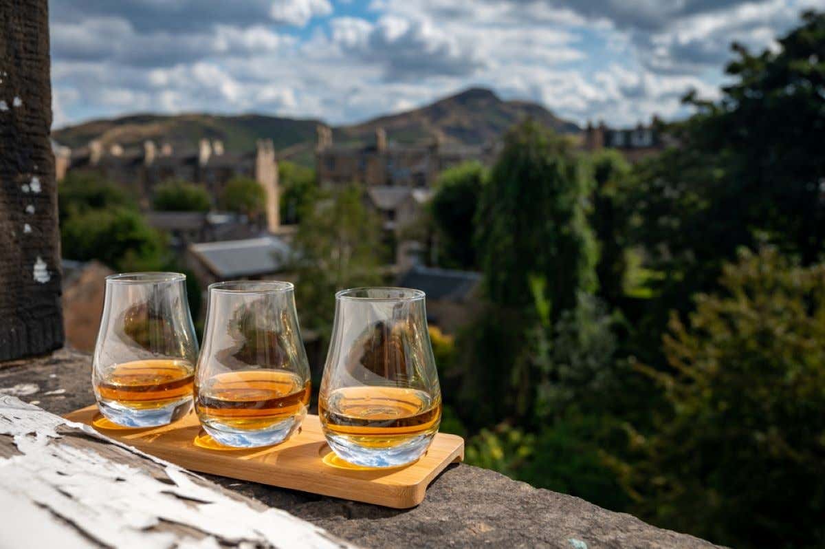 Three glasses of Scottish whisky on a wooden board with views of the city and hills of Edinburgh.