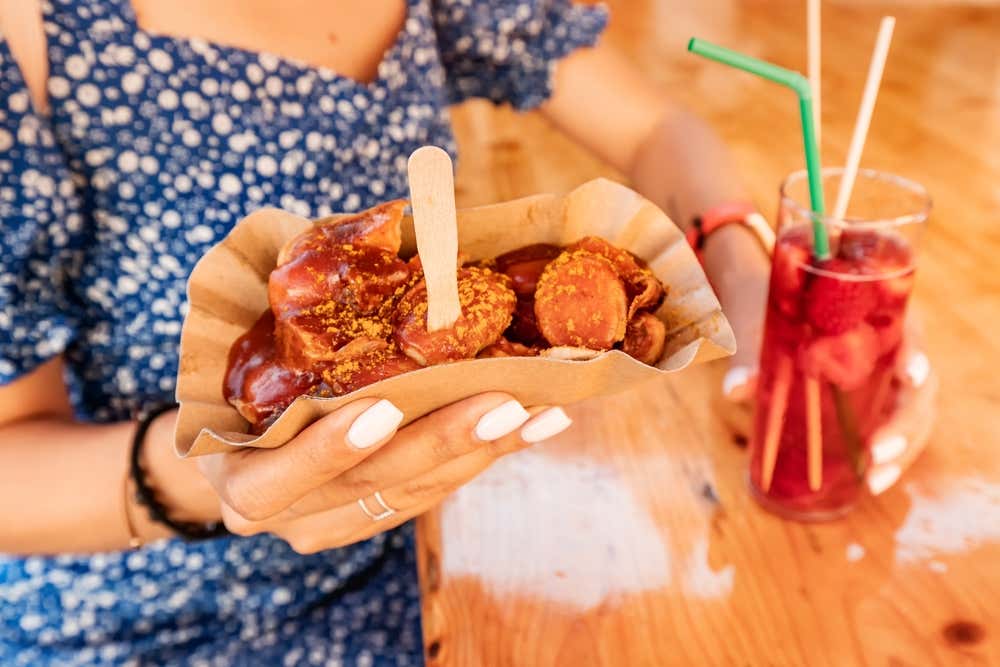 Manos sosteniendo una bandeja de currywurst picado junto a un vaso con bebida roja y pajita.
