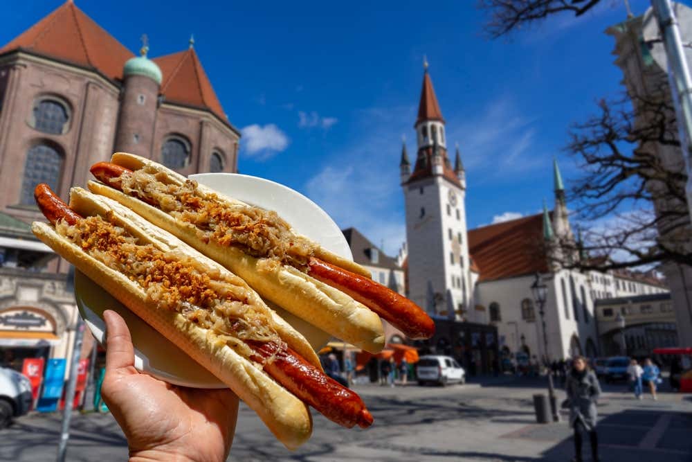 Una mano sujetando un perrito caliente en el Viktualienmarkt, y el ayuntamiento de Múnich al fondo