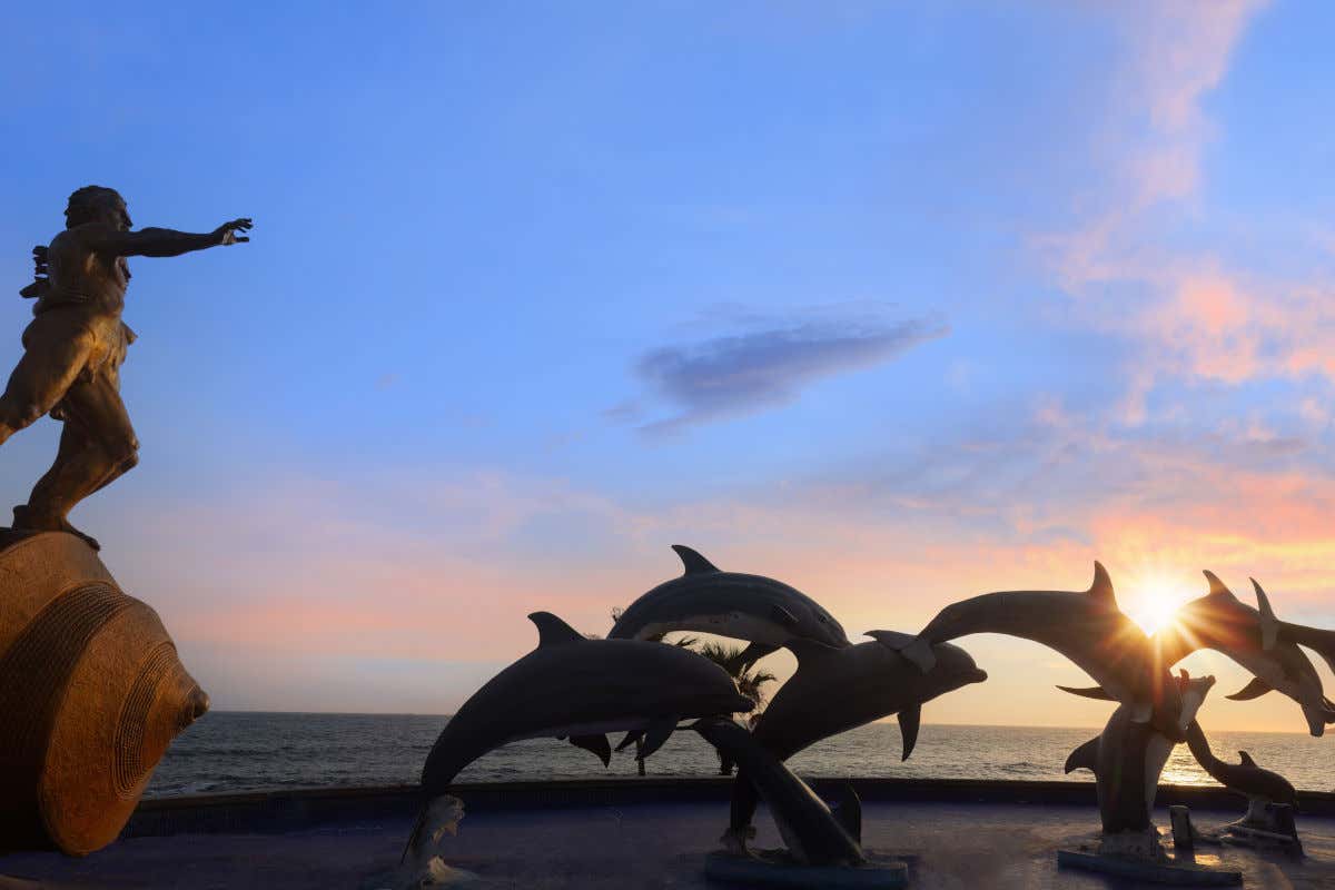 Escultura de un hombre y varios delfines junto al mar al atardecer