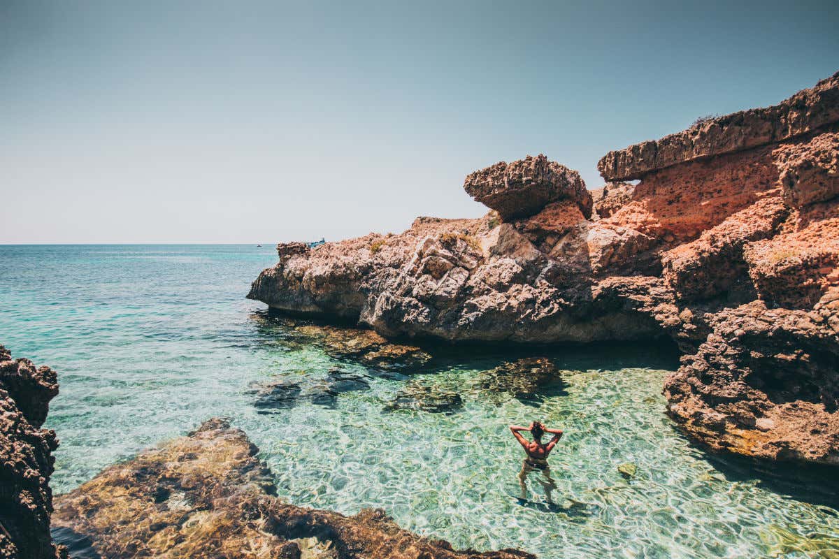 Una persona de espaldas a la cámara disfrutando de un baño en una cala de aguas transparentes y orillas rocosas