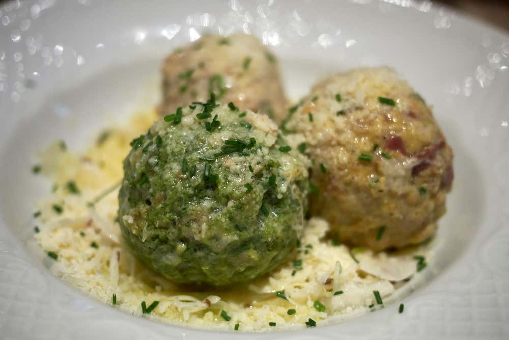 Tre grandi Knödel (canederli) austriaci, di cui uno verde brillante agli spinaci in primo piano e due più chiari con speck dietro, serviti in una fondina bianca su un letto di formaggio grattugiato e cosparsi di erba cipollina fresca tritata