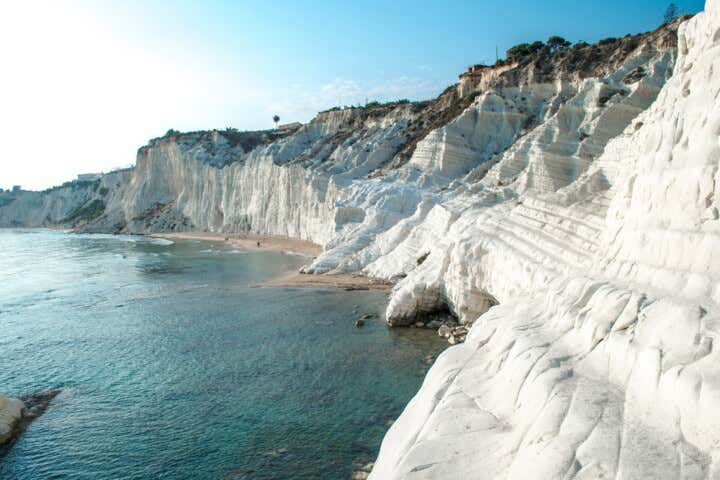 Veduta costiera della Scala dei Turchi in Sicilia, con l'imponente scogliera di marna bianca a forma di gradinata che scende fino al mare cristallino turchese, sotto un cielo azzurro e limpido con il sole alto a sinistra.