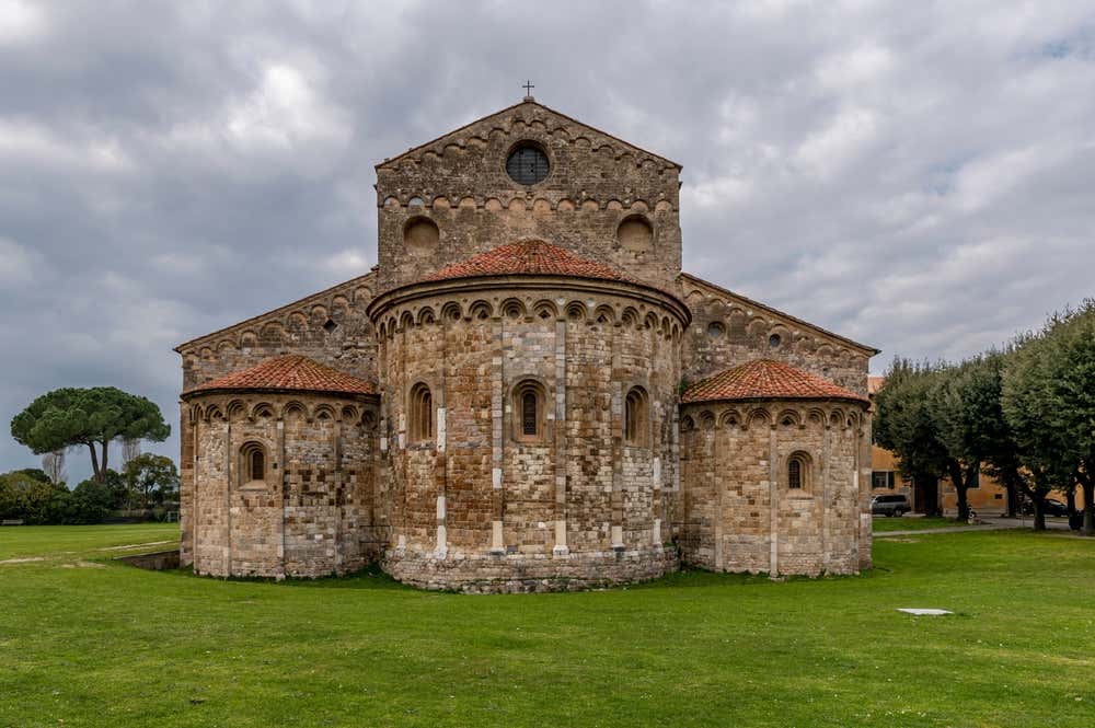 L’abside della basilica di San Piero a Grado, vicino a Pisa, in stile romanico pisano: tre absidi semicircolari in pietra con tetti di coppi rossi, decorate da archetti pensili, lesene e finestre a monofora. Sopra si vede la parte alta della chiesa con un oculo e una croce in cima, circondata da un prato verde e alberi sotto un cielo grigio e nuvoloso