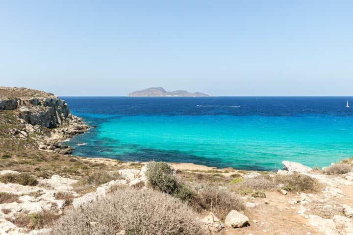 Veduta dall'alto della baia di Cala Rossa a Favignana: scogliera calcarea e arida in primo piano, acque trasparenti che sfumano dal turchese chiaro al blu intenso, con un'isola all'orizzonte sotto un cielo limpido
