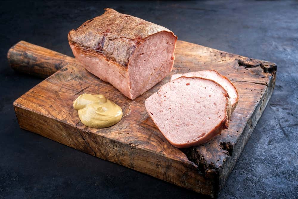 Un blocco di un polpettone di carne compatta, il Leberkäse, cotto al forno con crosta dorata appoggiato su un tagliere di legno rustico, con due fette spesse già tagliate in primo piano e una noce di senape al lato