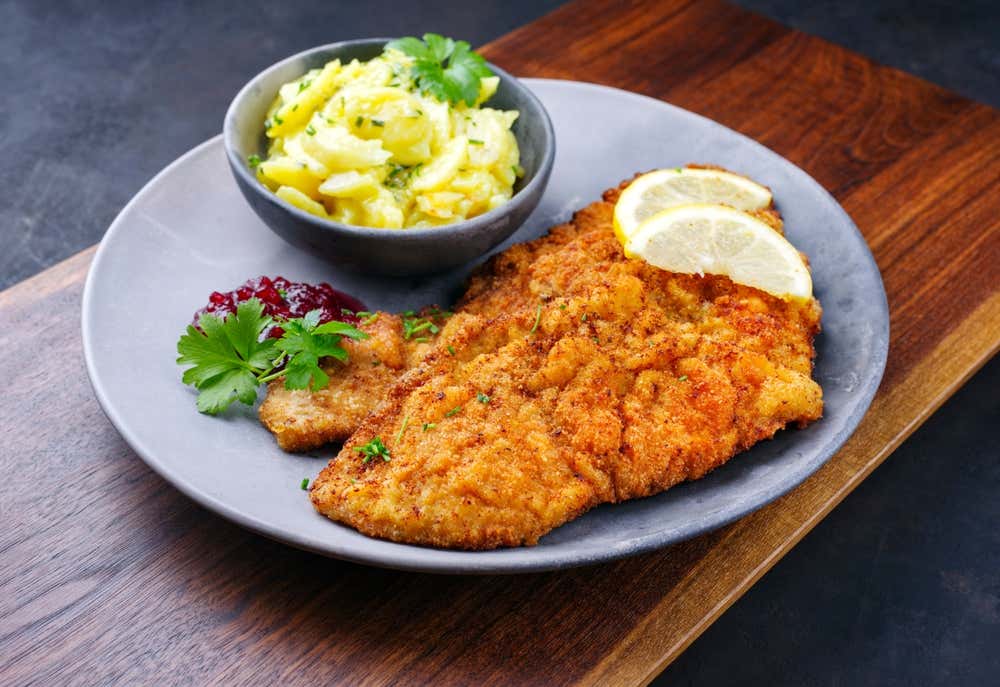 Una croccante cotoletta viennese dorata servita su un piatto grigio con due fette di limone sopra, accompagnata da un ciotolino di insalata di patate, confettura di mirtilli rossi e prezzemolo fresco, il tutto appoggiato su un tagliere di legno