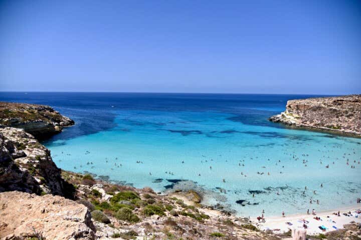 La spiaggia del coniglio a Lampedusa, vista dall'alto cinta come in un abbraccio da due scogliere, l'acqua è turchese e la sabbia bianca, in acqua si vedono alcuni bagnanti