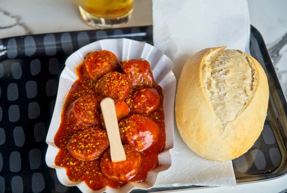 Un vassoio da chiosco con una vaschetta di carta contenente rondelle di salsiccia affogate in un'abbondante salsa rossa e spolverate di curry, con una forchettina di legno inserita; accanto, un panino croccante (Semmel) e un tovagliolo di carta
