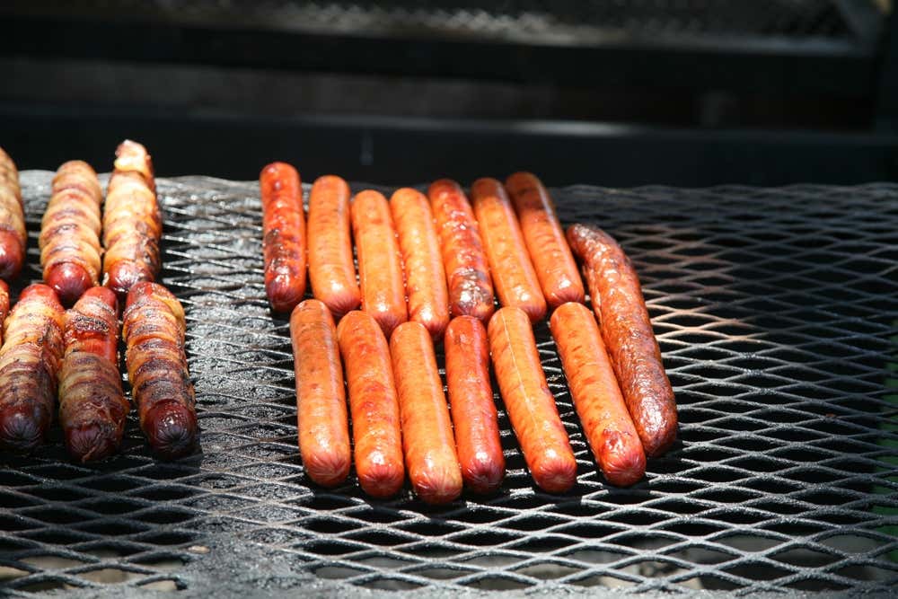 Una griglia in metallo piena di salsicce rosolate e dorate, disposte in fila; sulla sinistra alcune salsicce sono avvolte nella pancetta, mentre al centro e a destra ci sono dei classici Frankfurter allungati
