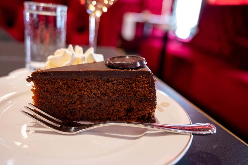 Una foto in primo piano di una fetta di Sacher Torte con panna montata e il sigillo di cioccolato dell'Hotel Sacher, servita su un piatto di porcellana bianca con bordo dorato e una forchetta d'argento, posizionata su un tavolo in un elegante caffè viennese. Sullo sfondo sfocato si intravedono lussuosi divani in velluto rosso e una calda illuminazione