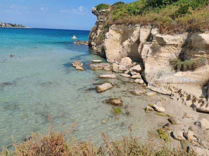 Uno scorcio ravvicinato della costa di Fontane Bianche, a Siracusa. In primo piano, l'acqua cristallina e bassissima sfuma dal trasparente al turchese, rivelando un fondale sabbioso con scogli arrotondati. A destra, una scogliera calcarea bianca e friabile, scolpita dall'erosione e sormontata da macchia mediterranea verde. In lontananza, si intravedono bagnanti in acqua, tra cui una persona su un materassino giallo, e una costa lontana sotto un cielo azzurro e sereno