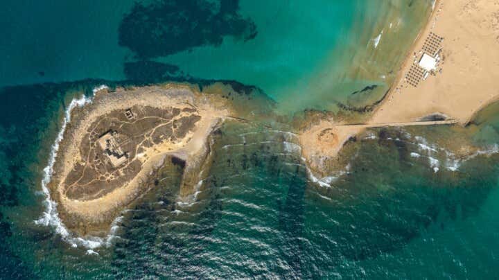 Fotografia aerea a piombo dell'Isola delle Correnti a Portopalo di Capo Passero. A sinistra, un isolotto roccioso e arido con le rovine di un vecchio faro è collegato alla terraferma da una sottile striscia di scogli artificiali e bassifondiSommersi. Il mare circostante sfuma dal verde turchese al blu profondo, con onde che si infrangono sui lati. A destra si stende una vasta spiaggia di sabbia dorata con uno stabilimento balneare