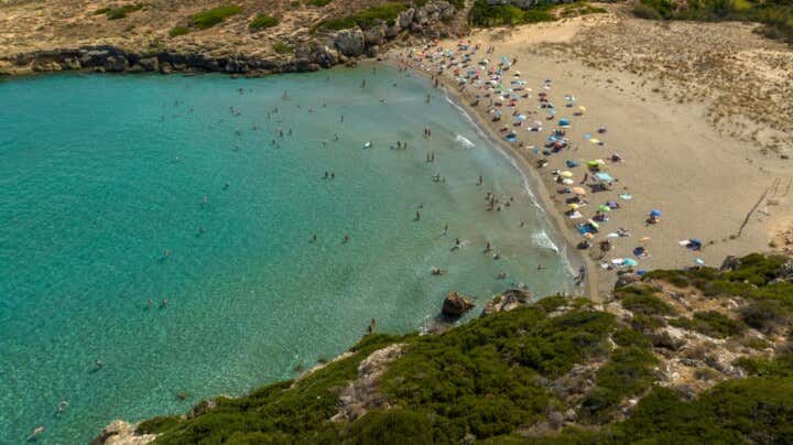 Veduta aerea della spiaggia di Calamosche in Sicilia. Una baia stretta e protetta tra scogliere, con acque cristalline color smeraldo popolate da bagnanti, che si infrangono su una spiaggia sabbiosa cosparsa di ombrelloni colorati