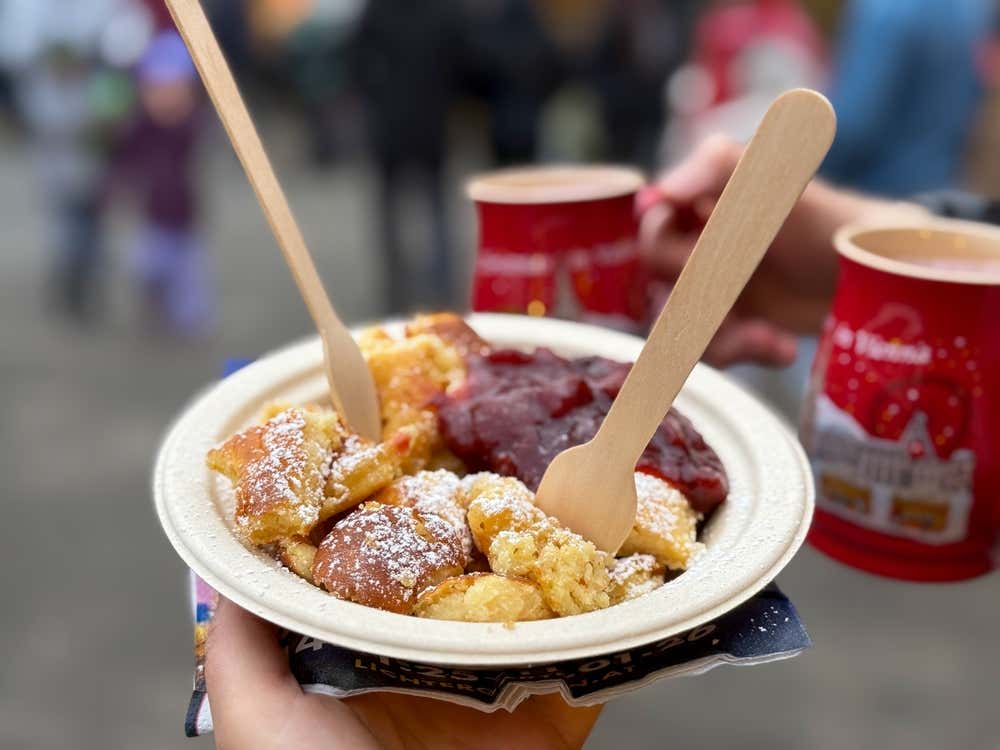 Una foto scattata a un mercatino di Natale affollato e sfocato, che mostra una mano in primo piano che regge una ciotola di carta riempita con Kaiserschmarrn (crêpe sminuzzate) cosparso di zucchero a velo e accompagnato da un mucchietto di marmellata di mirtilli rossi. Due forchette di legno sono inserite nel piatto. Sullo sfondo, si vedono due tazze di ceramica rossa per il vin brulé