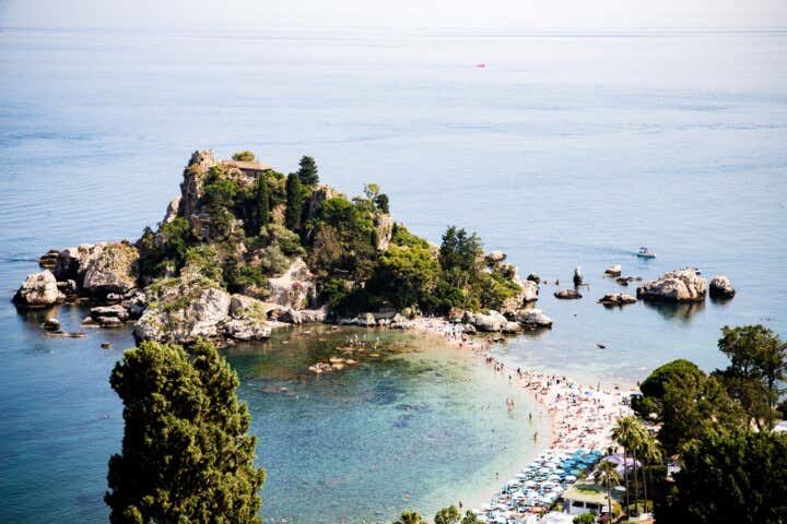 Una foto dall'alto di Isola Bella a Taormina, in Sicilia, che mostra una rigogliosa isola rocciosa collegata a una spiaggia sabbiosa da una stretta striscia di terra. L'isola è coperta da vegetazione verde scuro e presenta un edificio sulla sua sommità. La spiaggia è affollata di bagnanti e ombrelloni colorati. Le acque cristalline sono turchesi vicino alla riva e sfumano in un blu intenso più al largo, con alcune barche a vela visibili in lontananza
