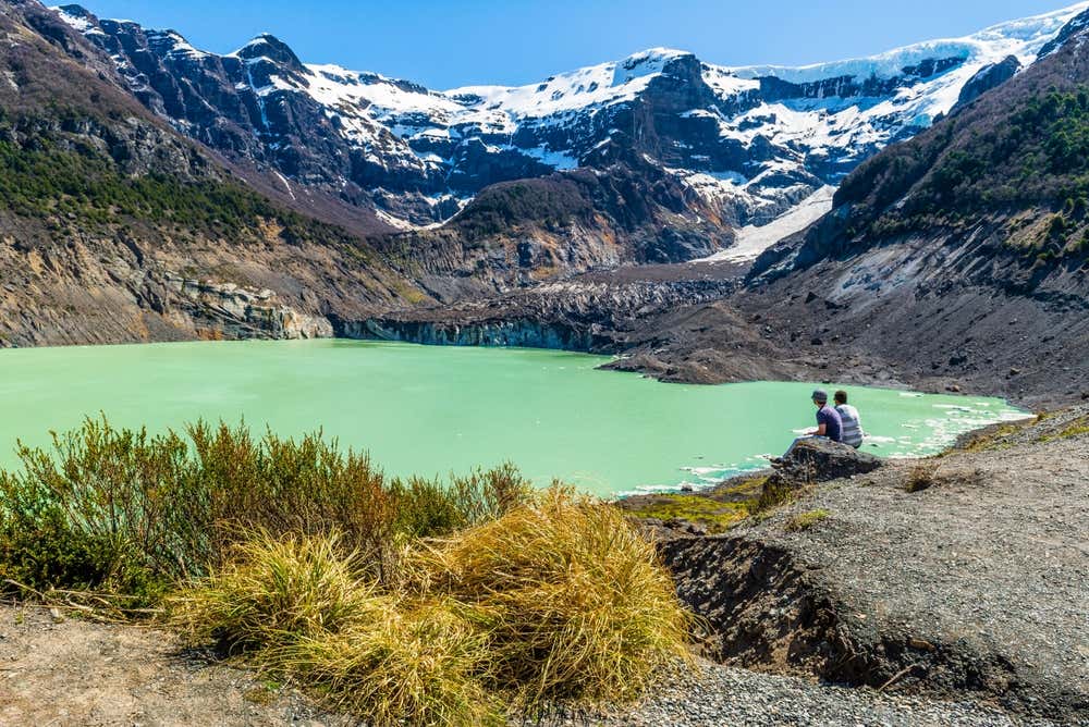 Dos personas sentadas frente a una laguna verde claro y montañas nevadas en el Ventisquero Negro