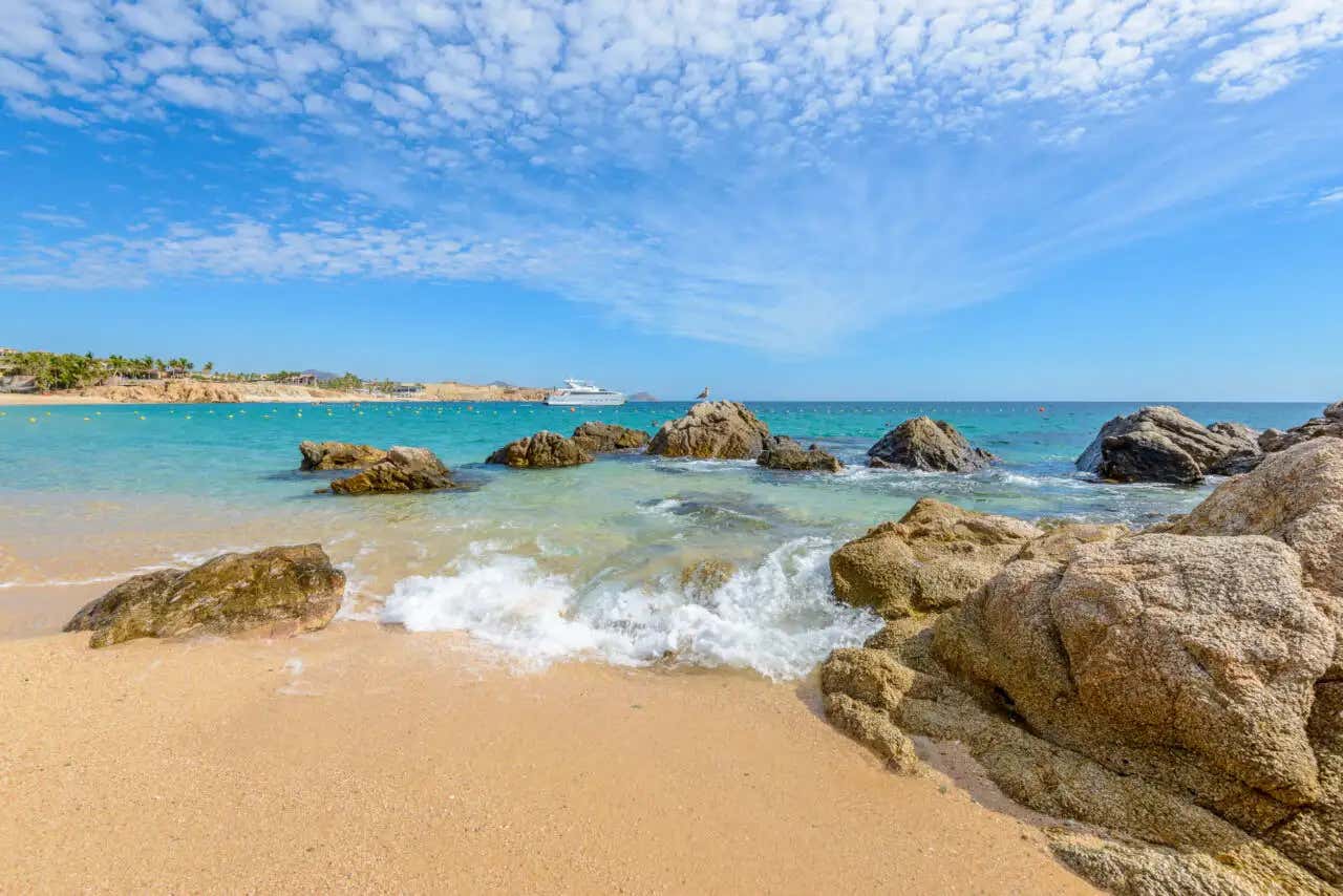 Una vista de una ensenada de playa arenosa y rocosa con agua turquesa cristalina. Grandes rocas están dispersas cerca de la orilla y en el agua. El cielo está lleno de nubes blancas y suaves. Una lancha a motor blanca es visible en la distancia