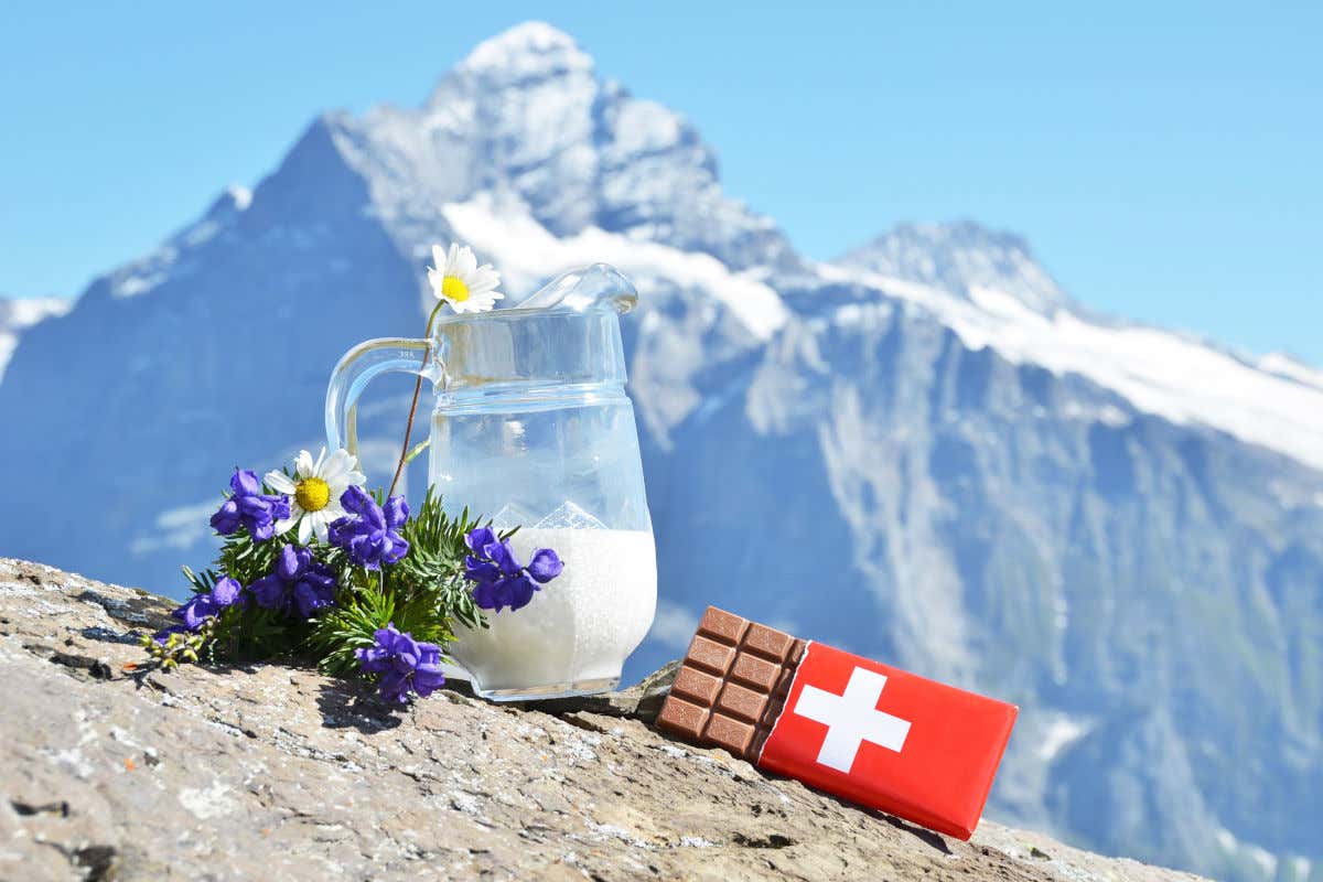 Una tableta de chocolate suizo junto a una jarra de leche y varias flores en una montaña nevada