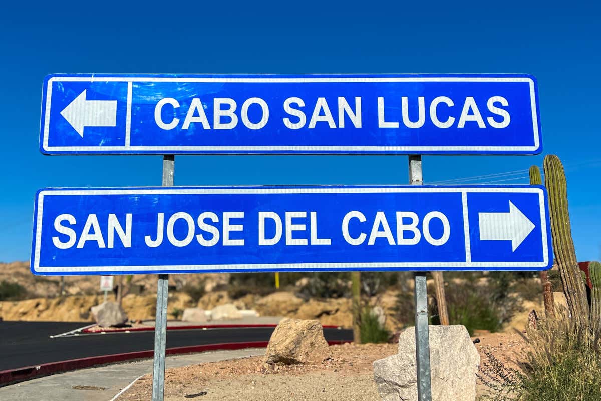 Un cartel de carretera de metal azul grande con letras blancas bajo un cielo azul brillante. La señal superior dice "CABO SAN LUCAS" con una flecha hacia la izquierda. La señal inferior dice "SAN JOSE DEL CABO" con una flecha hacia la derecha. El fondo es un paisaje desértico con cactus y colinas bajas