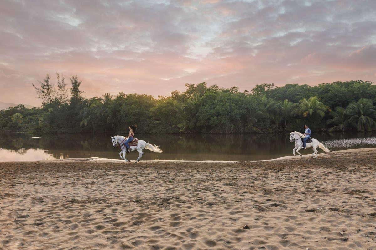 Dos jinetes cabalgando dos caballos blancos en una playa al atardecer